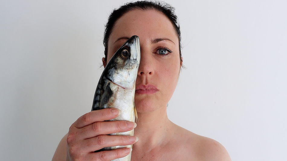 A person is holding a Mackerel fish up to their face, you can see their head with dark hair and the top of their shoulders.